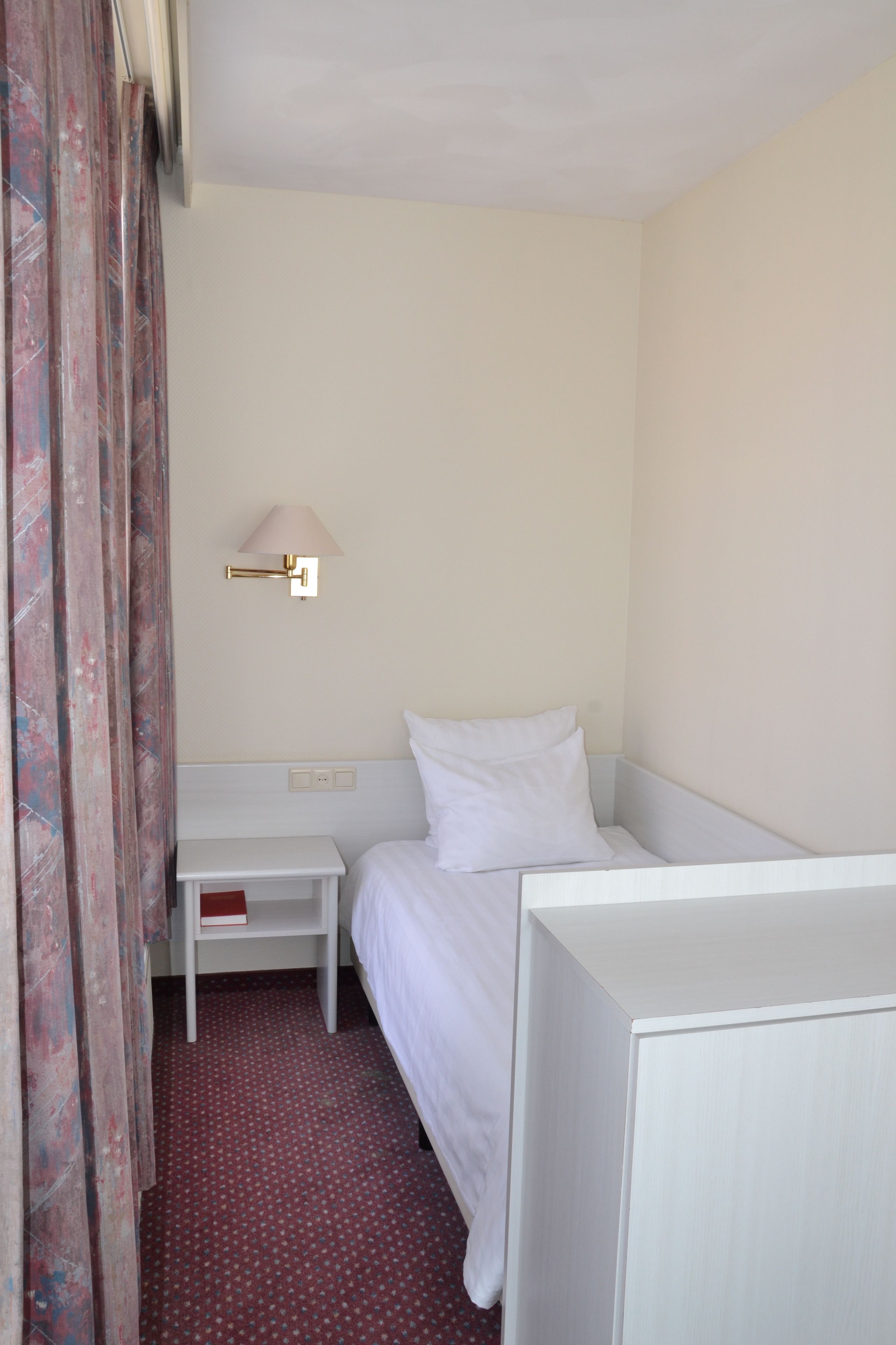Habitación de hotel con una cama individual, una mesita de noche, una lámpara y una cortina.