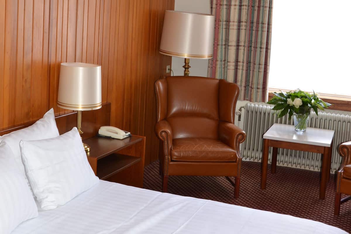 Een hotelkamer met een groot bed, een wandpaneel van hout, een bruine leren fauteuil, een klein tafeltje met een lamp en een raam met gordijnen.