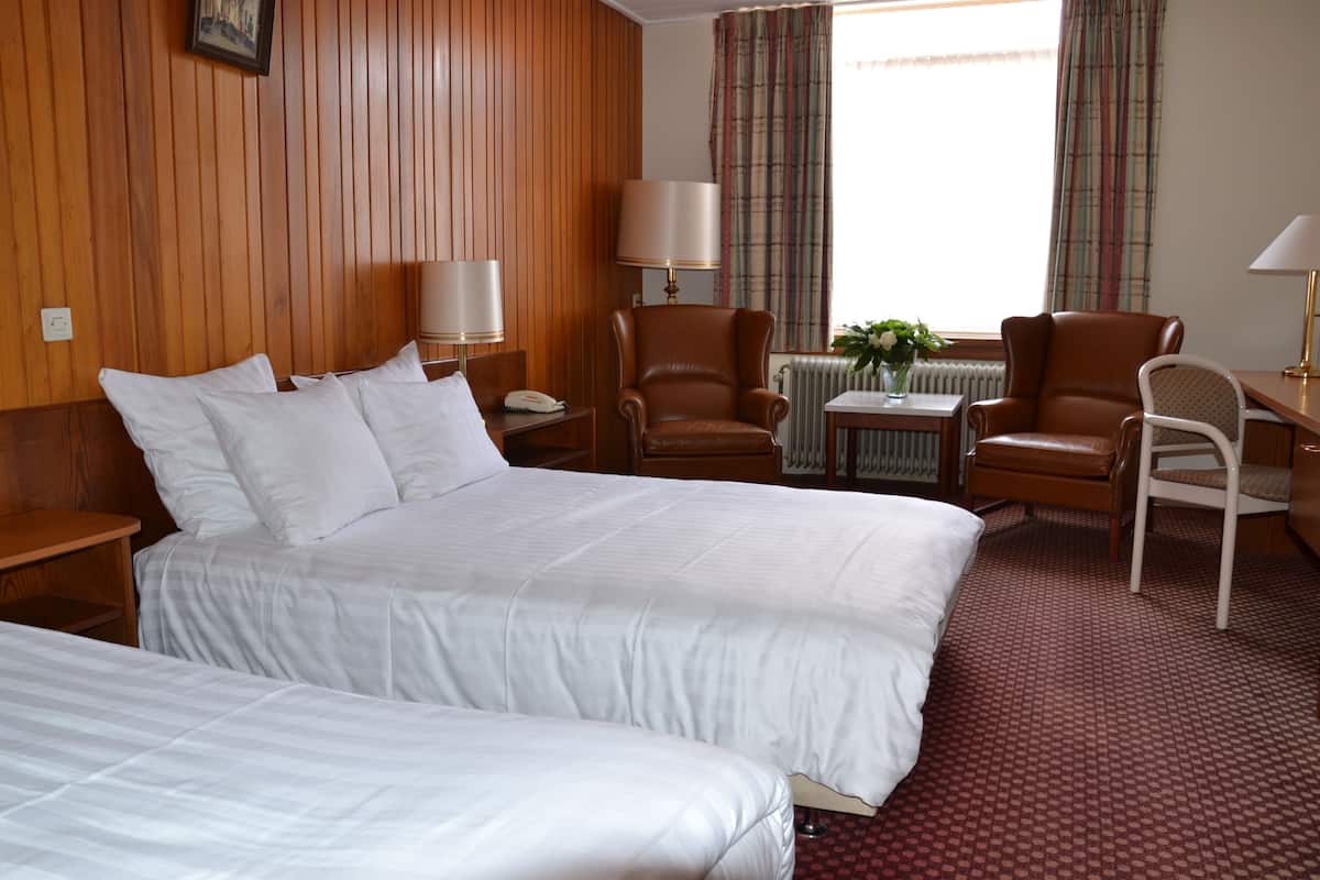 Een hotelkamer met twee bedden, houten lambrisering, een tafel met lamp, een stoel en een raam met gordijnen.