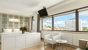 A modern hotel room with a large window, a flat-screen TV, a white sofa, a small round table, and a decorative vase with flowers. - NH Amsterdam Zuid (Amsterdam)