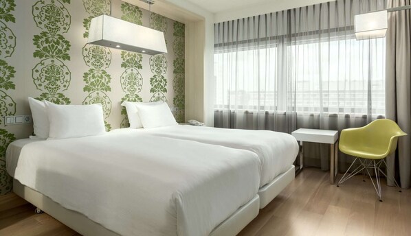 Quarto de hotel com duas camas, uma escrivaninha, uma cadeira e uma janela grande com cortinas.