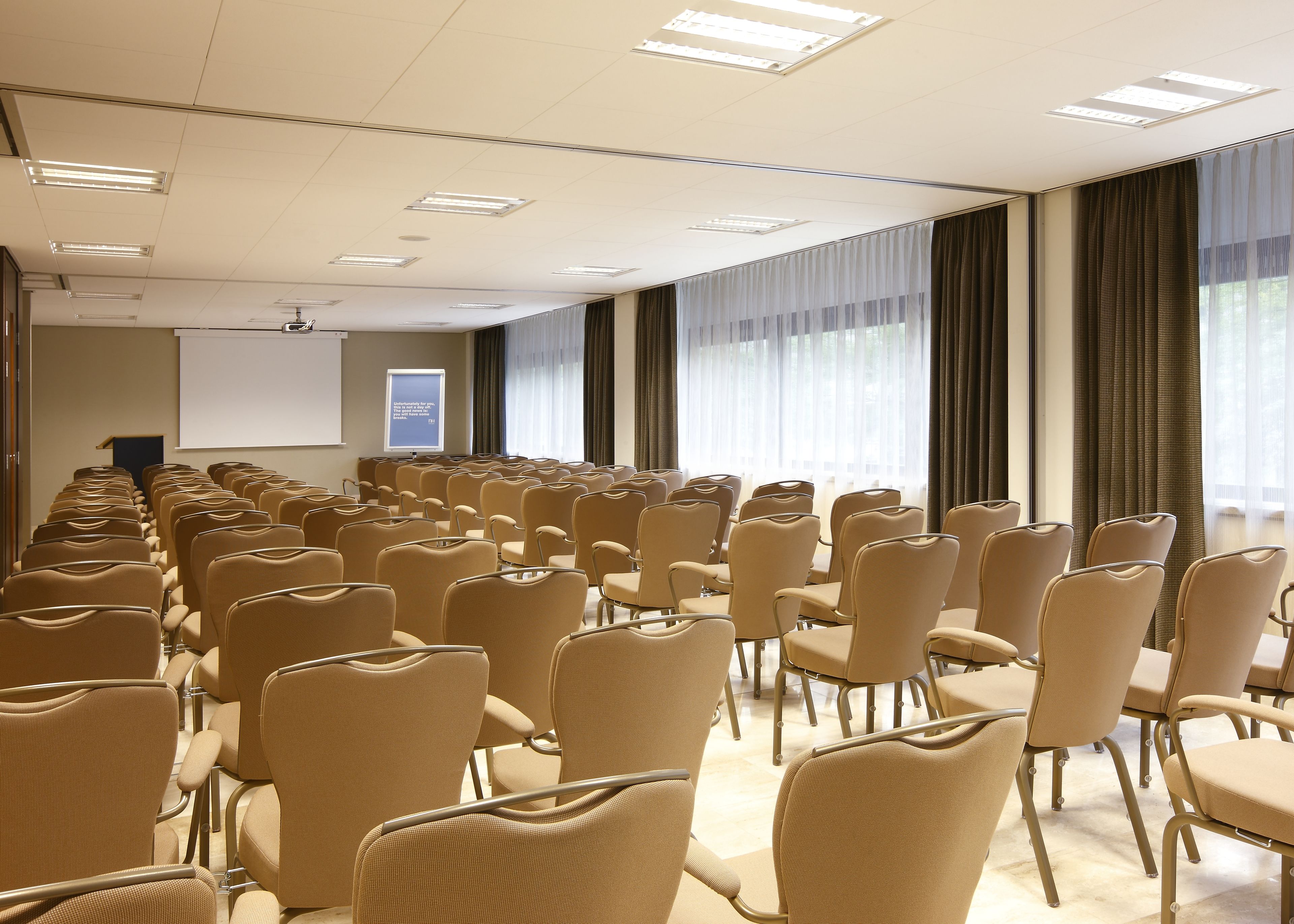 Un salón de conferencias con filas de sillas color beige, una pantalla de proyector y ventanas grandes con cortinas.