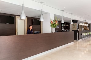 Un área de recepción moderna con mostrador de madera, iluminación colgante, una planta en maceta y una zona de bar con taburetes.