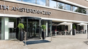 The exterior of NH Amsterdam ZUID hotel with a glass entrance, outdoor seating area, and potted plants. - NH Amsterdam Zuid (Amsterdam)
