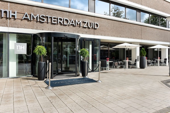 Exterior del hotel NH Amsterdam ZUID con entrada de cristal, zona de estar al aire libre y plantas en macetas.
