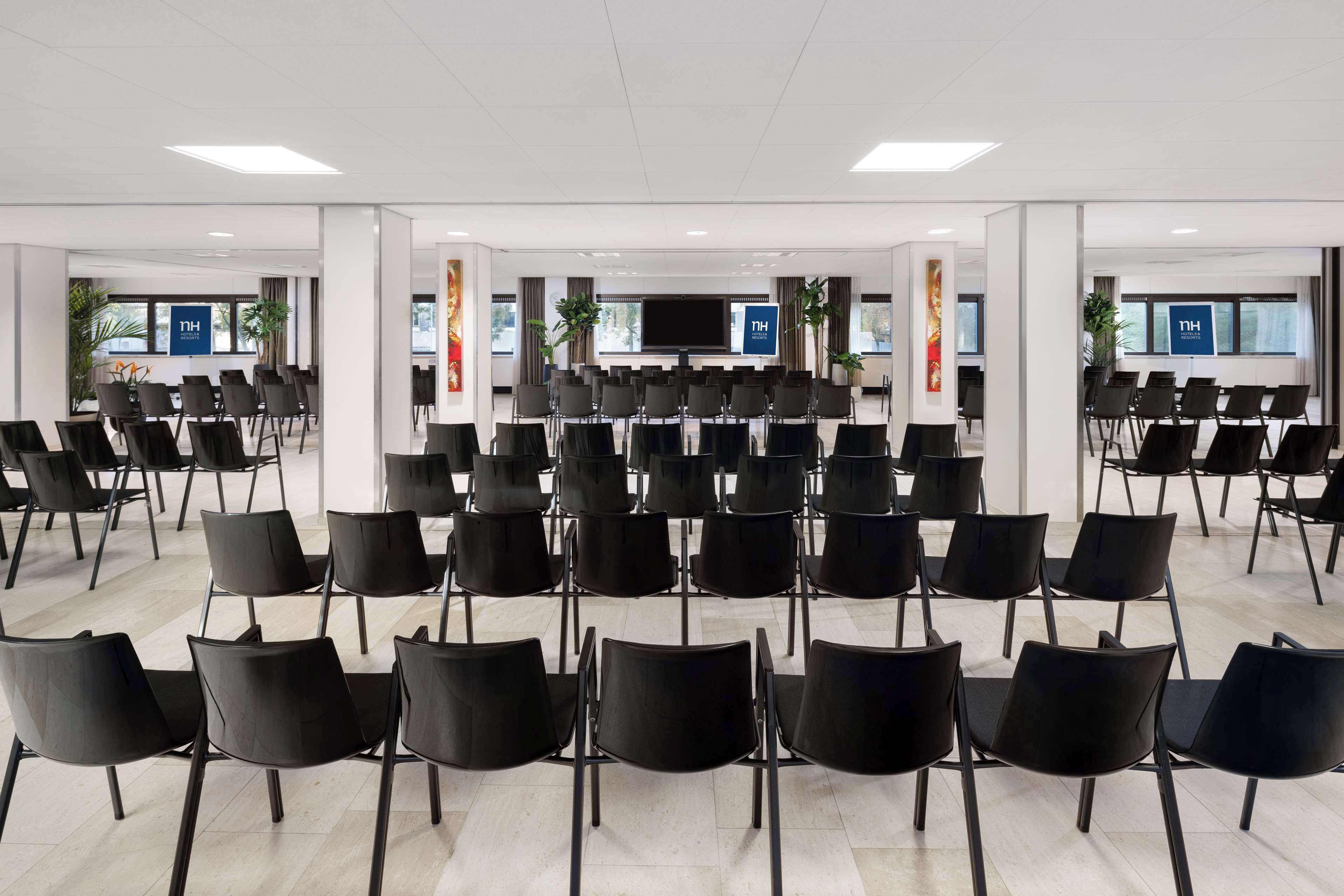 Een vergaderzaal met rijen zwarte stoelen, een groot scherm en potplanten.