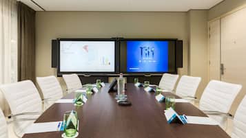 Una sala de conferencias con una mesa larga, sillas blancas, una pantalla de proyector que muestra gráficos y una pantalla con la marca del hotel.