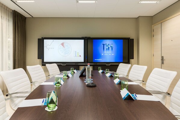 A conference room with a long table, white chairs, a projector screen displaying graphs, and a screen with hotel branding.