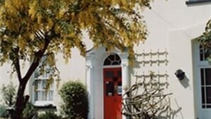 Un edificio blanco con puerta roja, un pequeño porche y un árbol de hojas amarillas al frente.