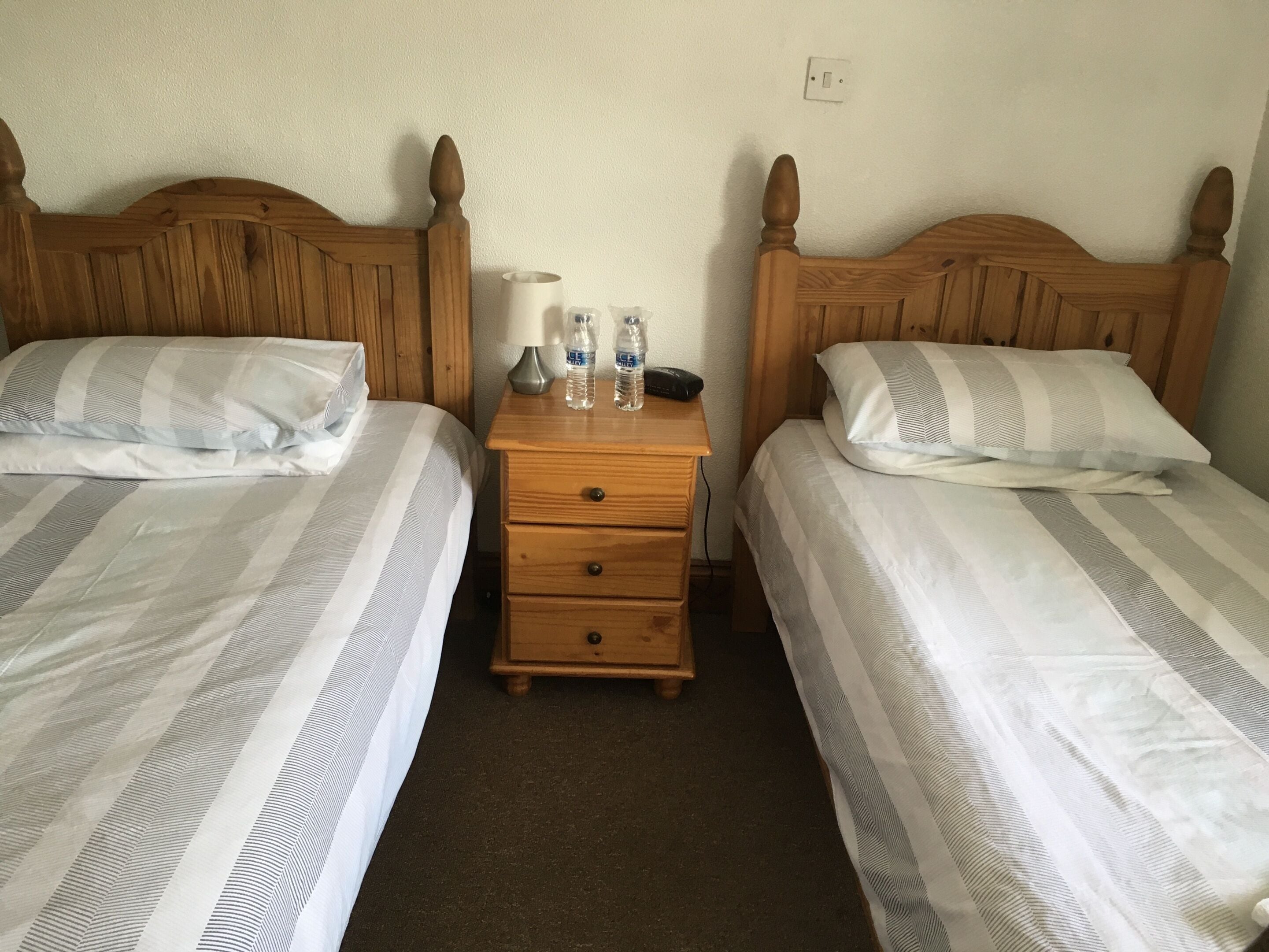 A double room with two beds, each with striped bedding and wooden headboards. There is a wooden nightstand between the beds with a lamp and two bottles of water.