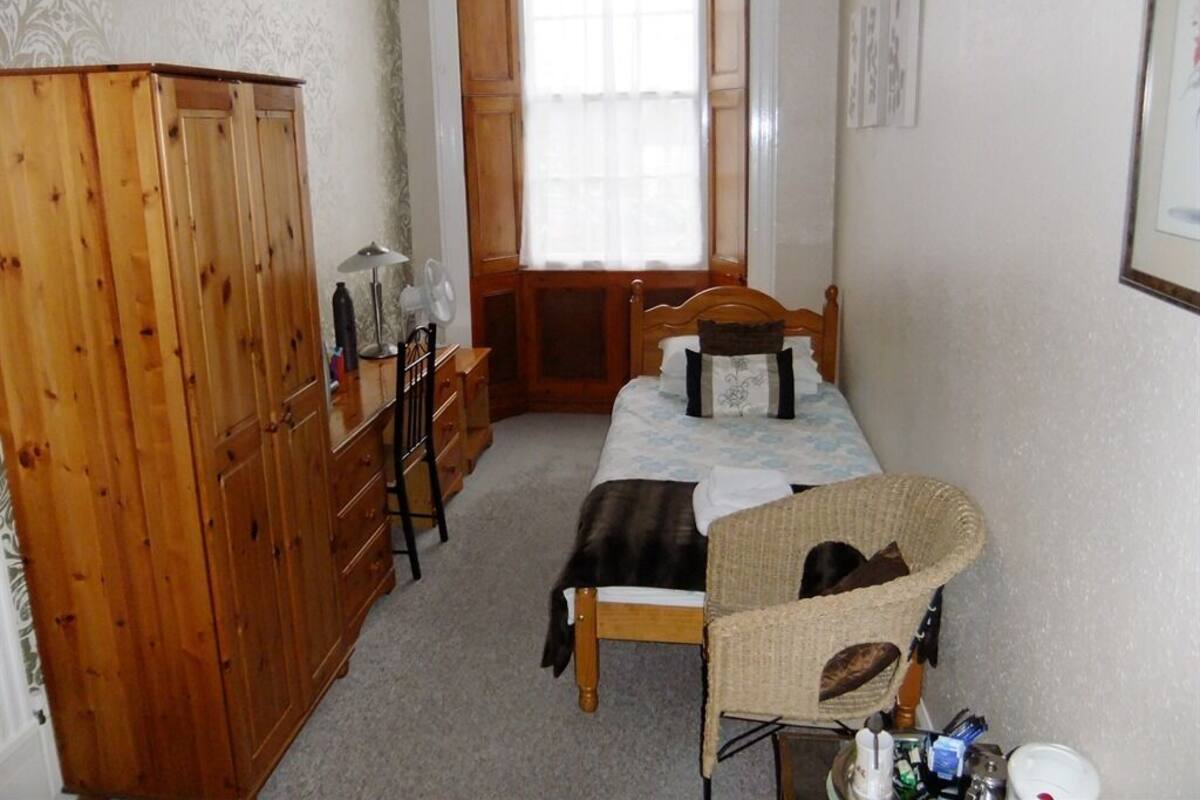 A bedroom with a wooden wardrobe, a bed with a headboard, a chair, and a window with curtains.