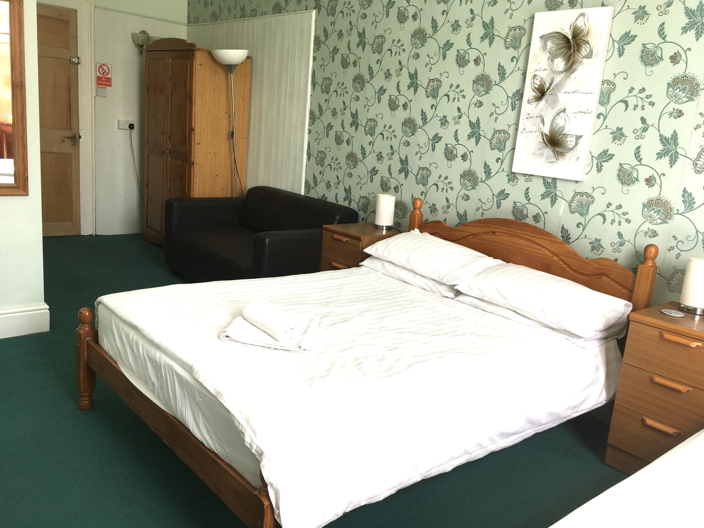 A neatly made bed with white linens, a wooden headboard, and a bedside table with a lamp. A black leather chair is positioned against the wall, and a wooden wardrobe is visible in the background.