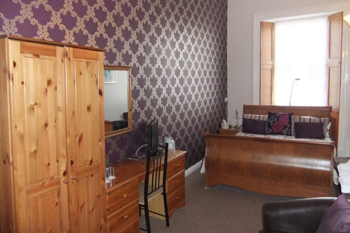 A bedroom with a wooden wardrobe, a desk with a chair, a bed with a headboard, and a window with curtains.
