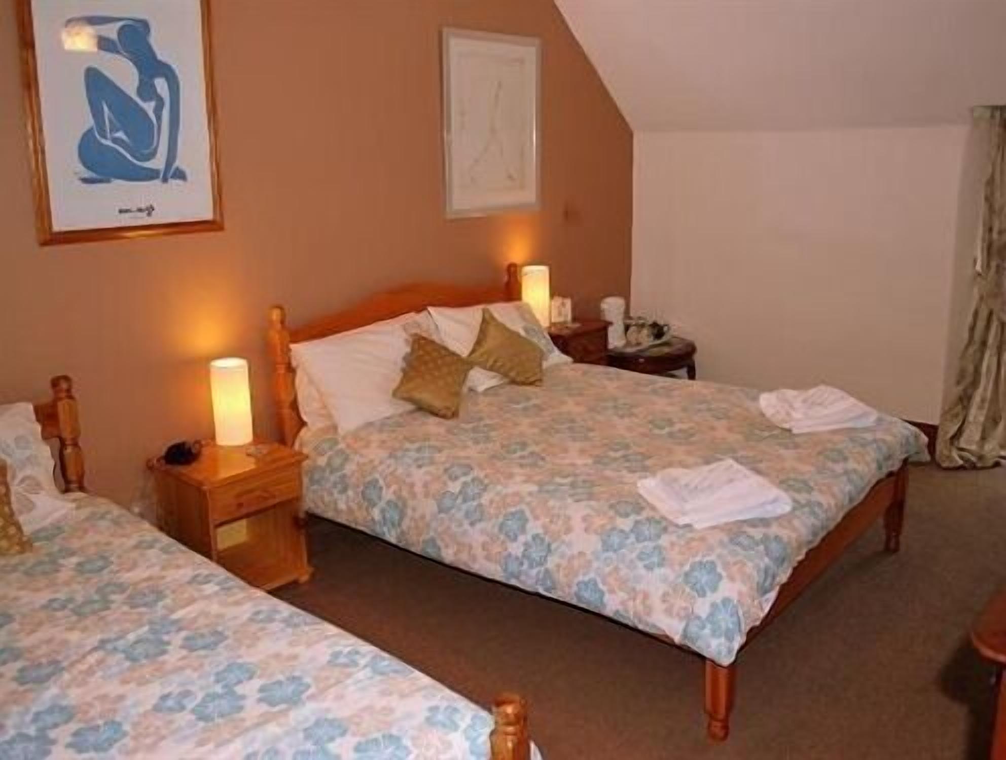 A bedroom with two beds, each with a floral bedspread and wooden headboards. There are bedside lamps, a nightstand, and a framed picture on the wall.