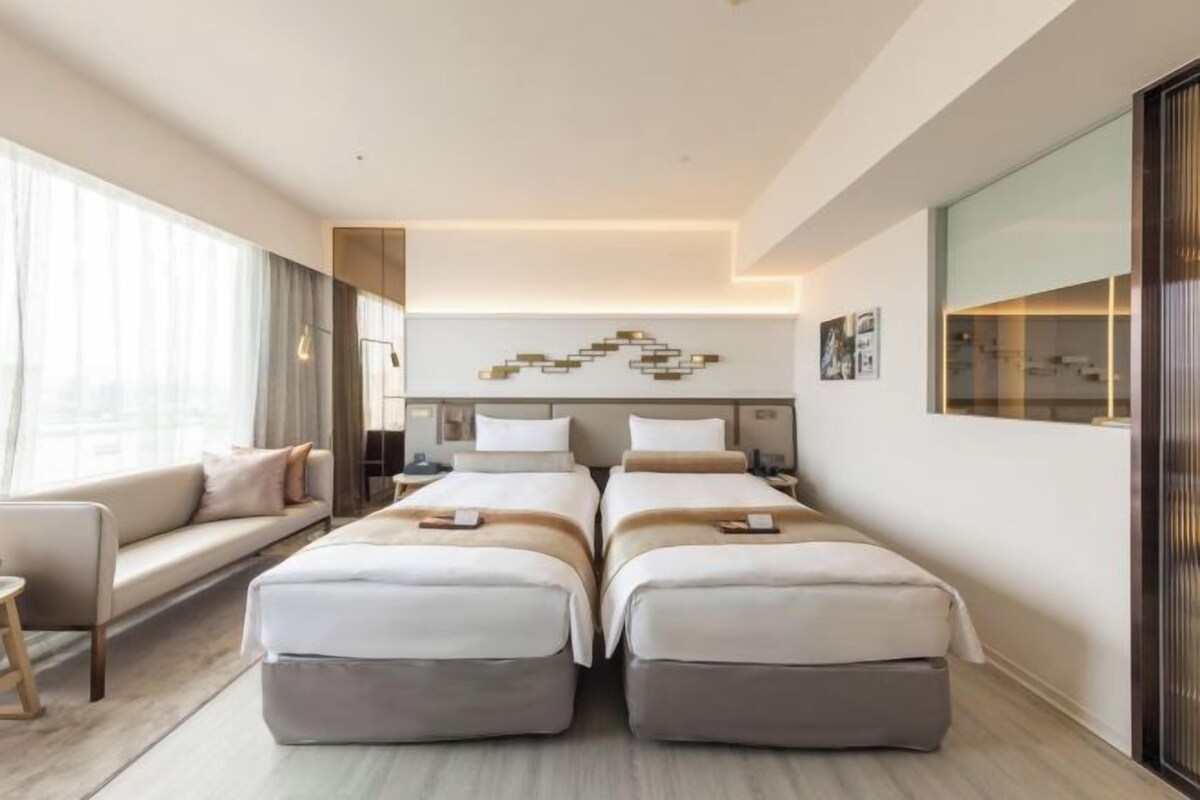 A modern hotel room with a sofa, two beds, a desk, and a large window.