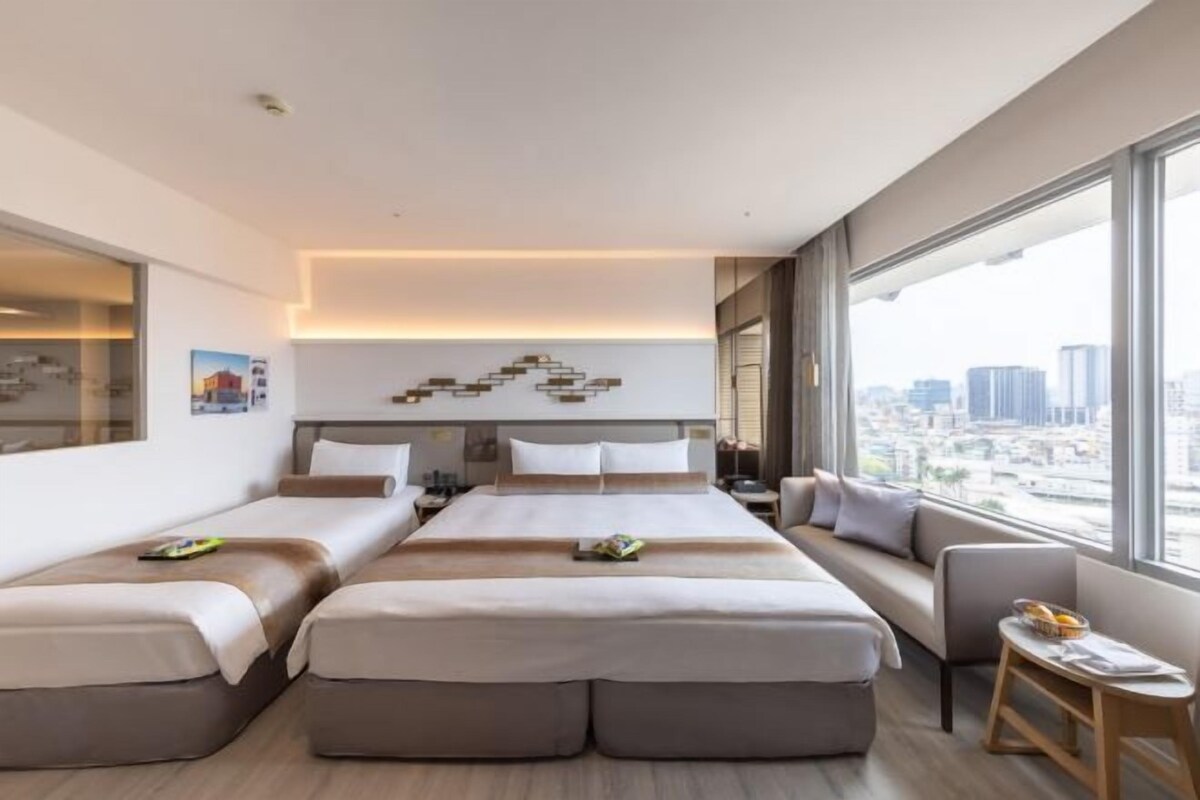 A modern hotel room with a large bed, a sofa, a small table, and a view of the city.