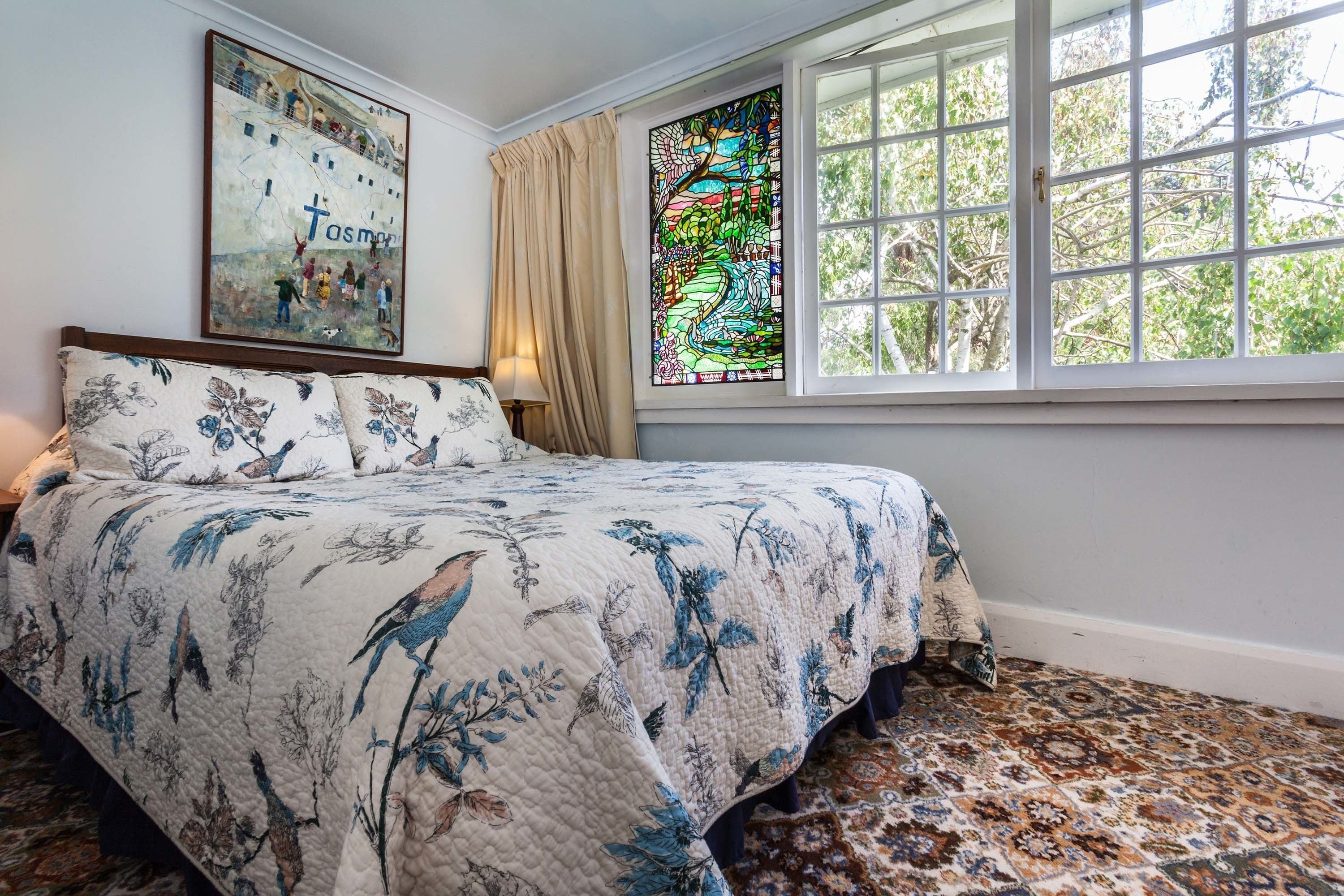 A bedroom with a bed, floral bedding, a stained glass window, and a view of trees.