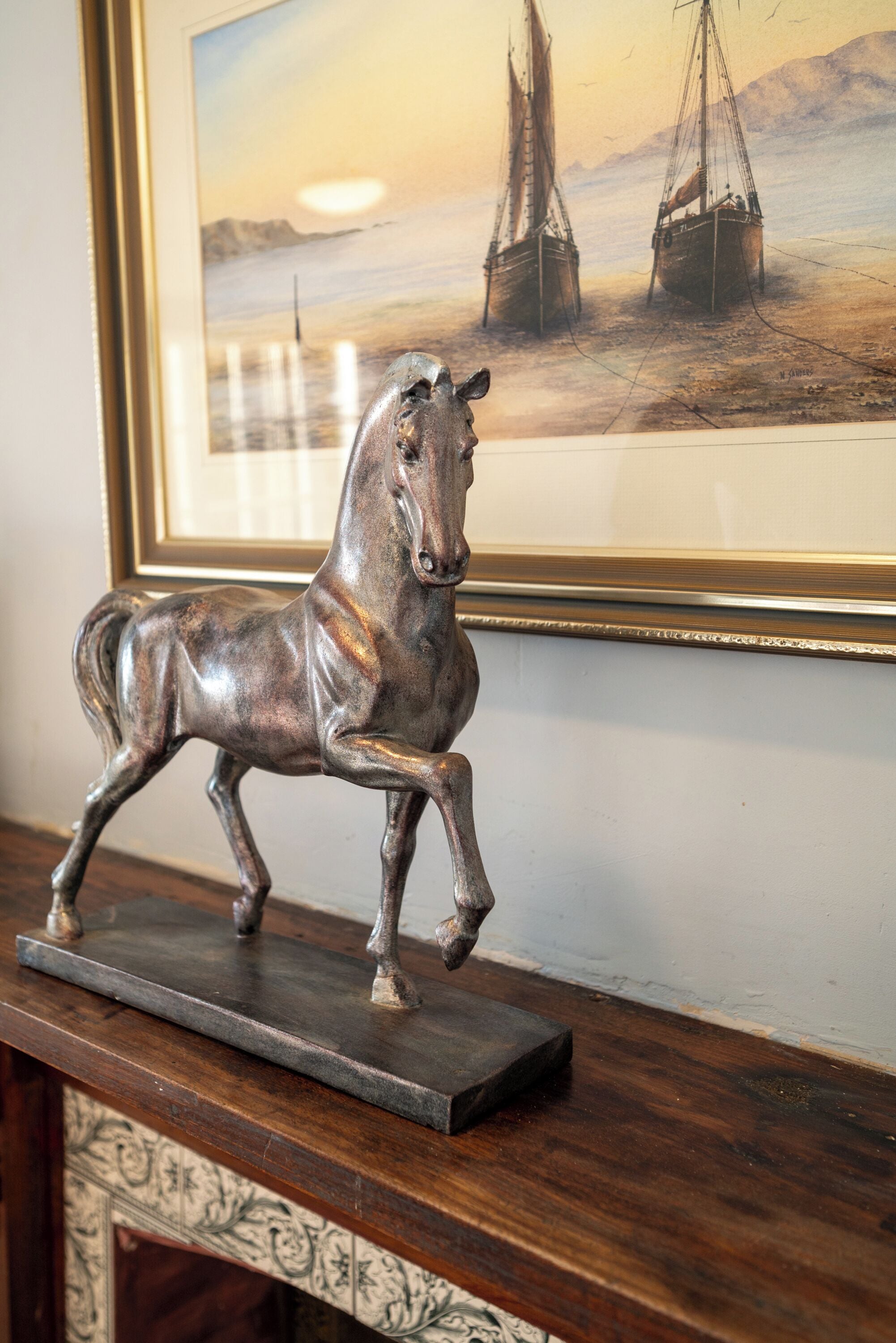 Une sculpture équestre en bronze, posée sur un socle en bois foncé, avec, en arrière-plan, un tableau encadré représentant des voiliers.