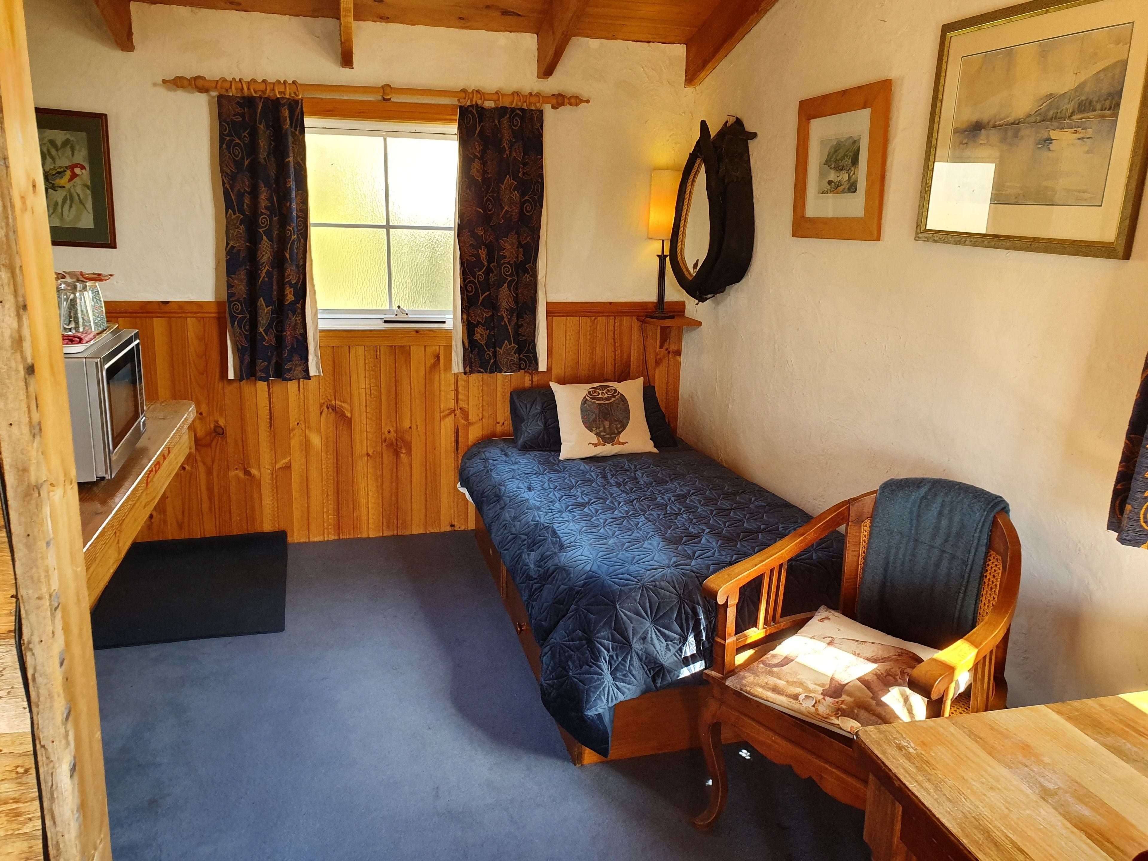A compact room with a bed, a wooden desk, a chair, a window with curtains, and a wall-mounted mirror.