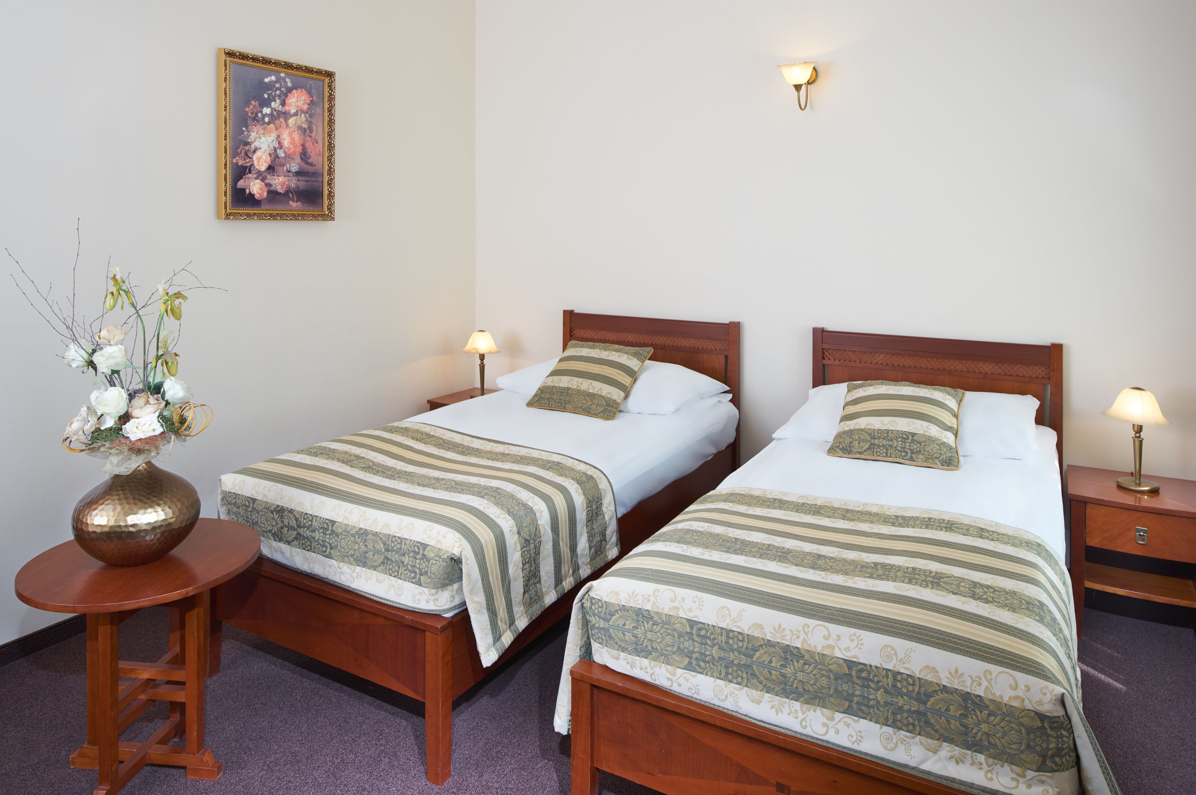 A hotel room with two single beds, a round wooden nightstand, a small table with a vase of flowers, and a framed floral painting on the wall.