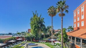 A swimming pool with lounge chairs and palm trees, surrounded by a building with a red facade and a tiled roof.