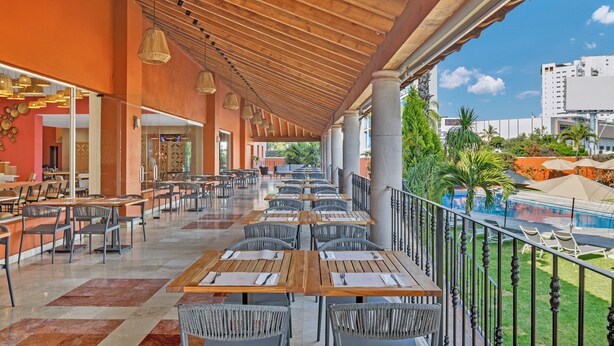 An outdoor dining area with wooden tables and chairs, a pool in the background, and a view of a cityscape.
