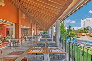 Un espacio para comer al aire libre con mesas y sillas de madera, una piscina al fondo y vistas a la ciudad.