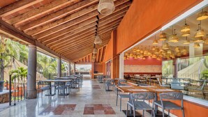 A spacious outdoor dining area with multiple tables and chairs, a wooden ceiling, and large windows offering views of the surrounding landscape.