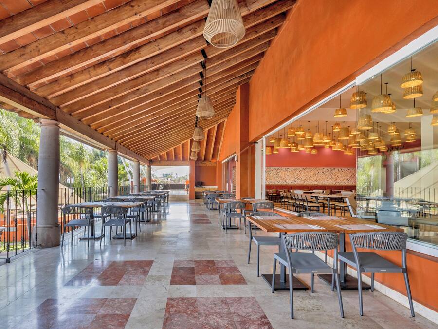 Un amplio espacio de comedor al aire libre con múltiples mesas y sillas, techo de madera y ventanas grandes que ofrecen vistas del paisaje circundante.