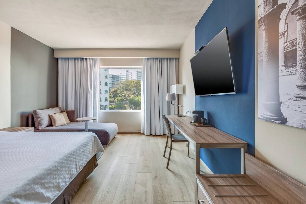 Habitación de hotel con cama, escritorio, silla, televisión y ventana con cortinas.