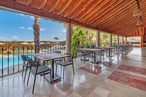 Un área de comedor al aire libre techada, con mesas y sillas, una piscina y palmeras.