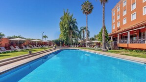 A swimming pool with lounge chairs and palm trees, surrounded by a building with a red facade.