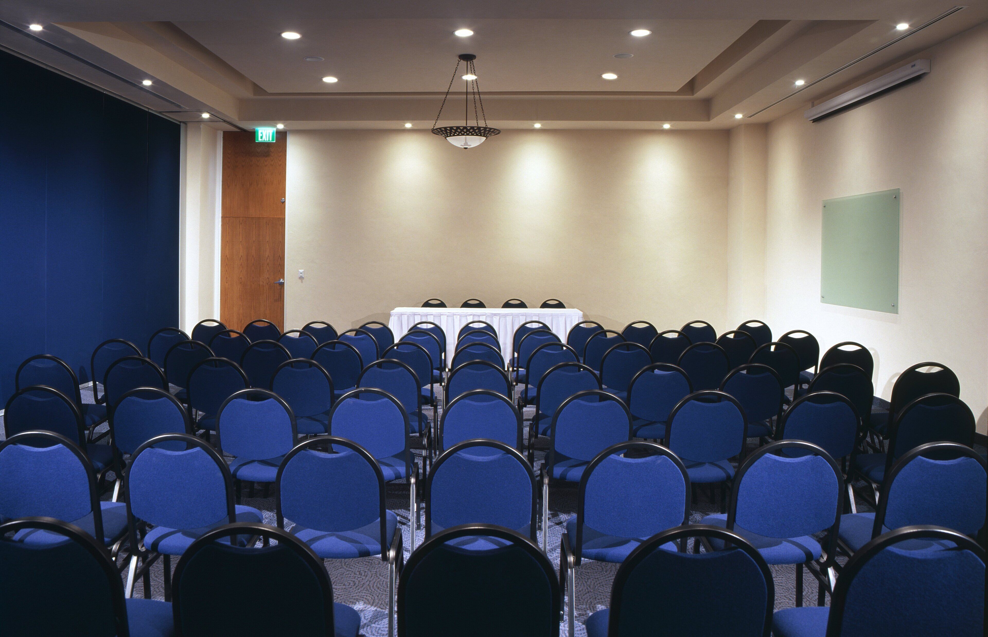 Konferenssihuone, jossa on riveissä sinisiä tuoleja, valkoisella pöytäliinalla peitetty pöytä ja kattoon asennettu projektori.