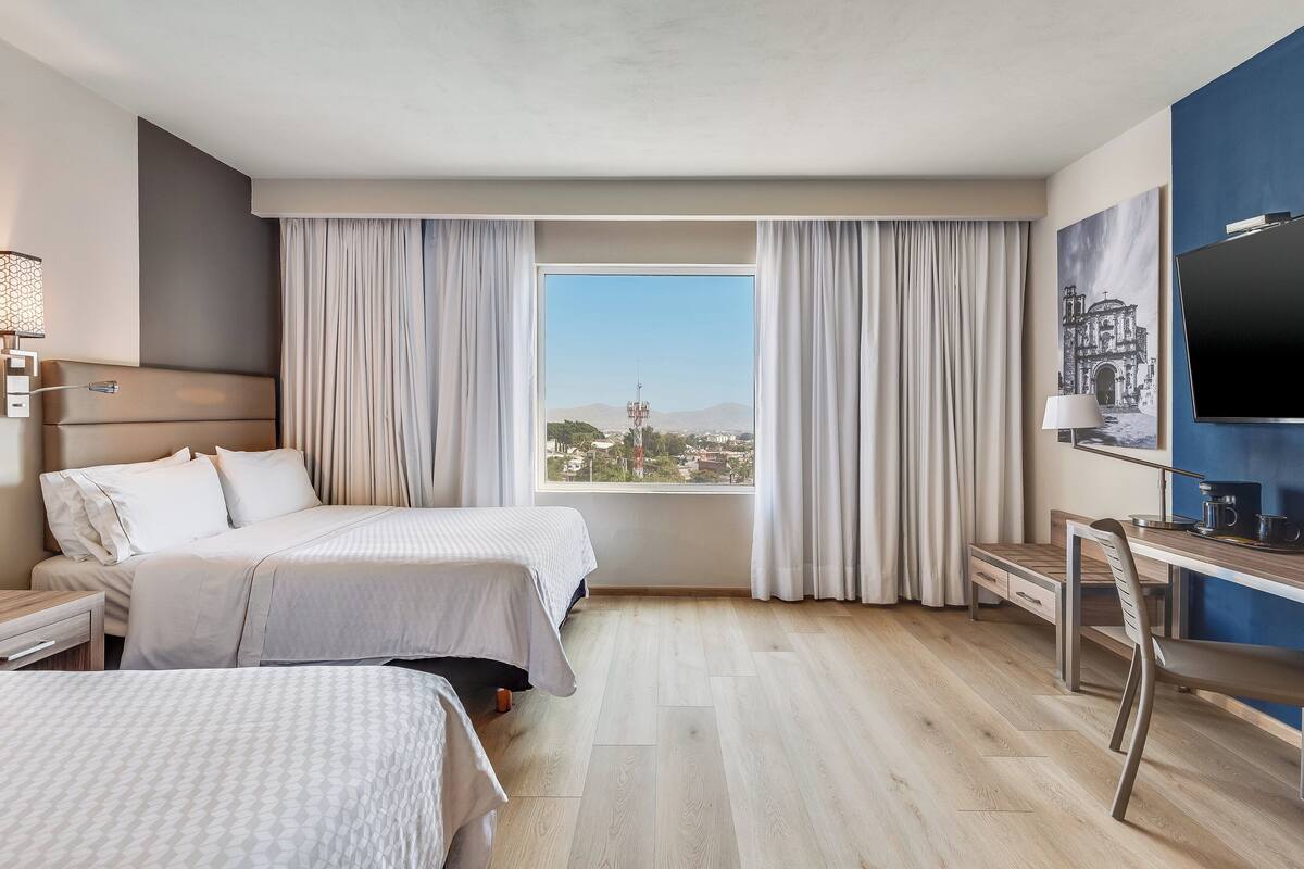 Habitación de hotel con dos camas, un escritorio, una silla, un televisor y un amplio ventanal con cortinas.
