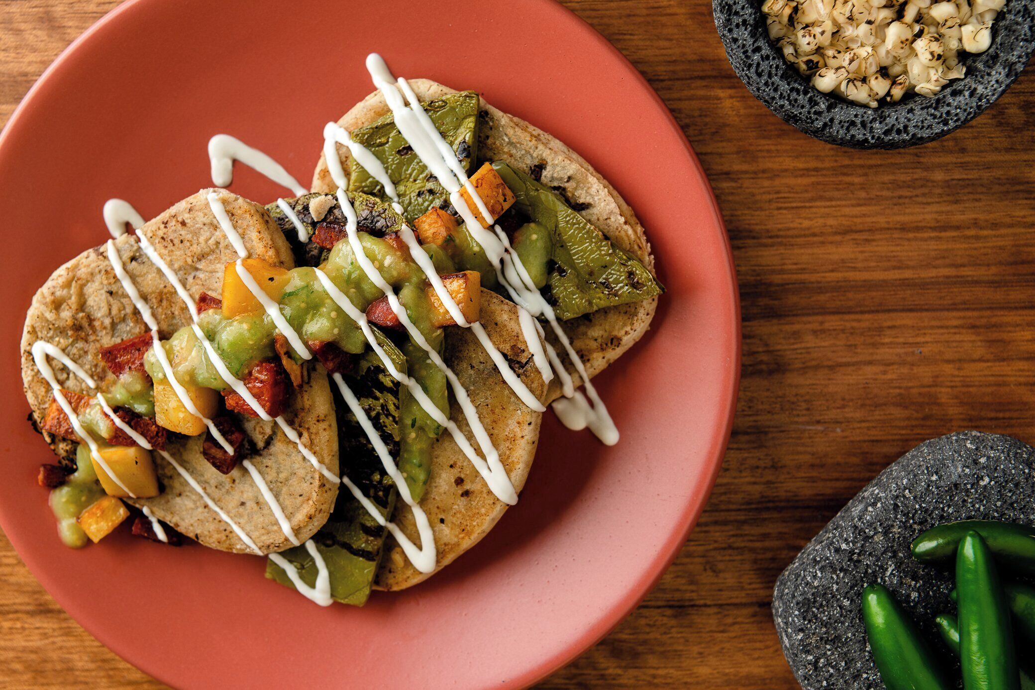 Un plato de tacos con diversos ingredientes y una salsa blanca, acompañado de un cuenco de palomitas de maíz y un mortero con chiles verdes.