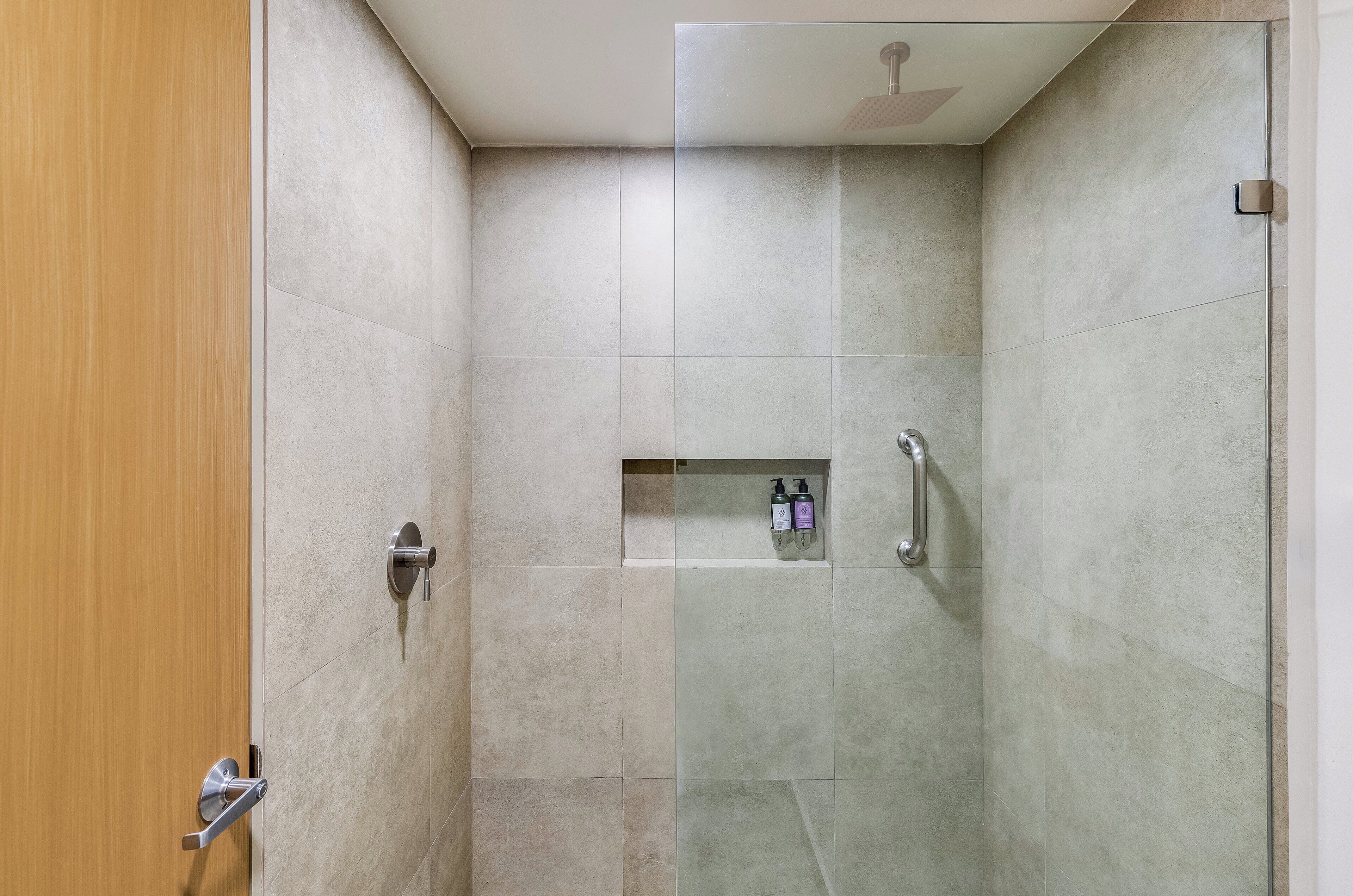 Ducha moderna con puerta de vidrio, estante empotrado con artículos de aseo y ducha suspendida.