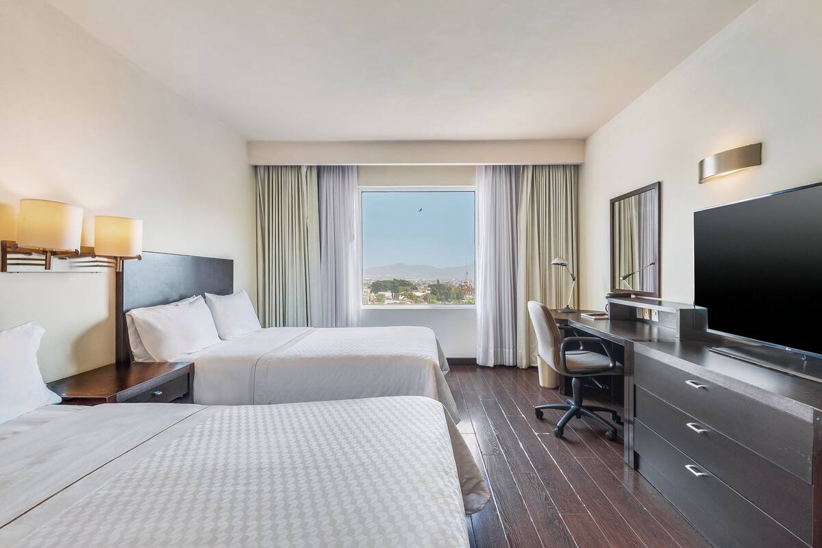 Habitación de hotel con dos camas, un escritorio, un televisor y un ventanal grande con cortinas.