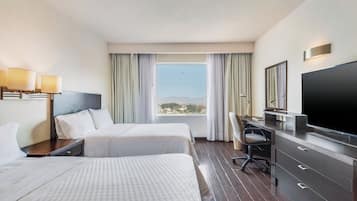 Habitación de hotel con dos camas, un escritorio, un televisor y un ventanal grande con cortinas.