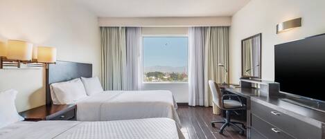 Habitación de hotel con dos camas, un escritorio, un televisor y un ventanal grande con cortinas.