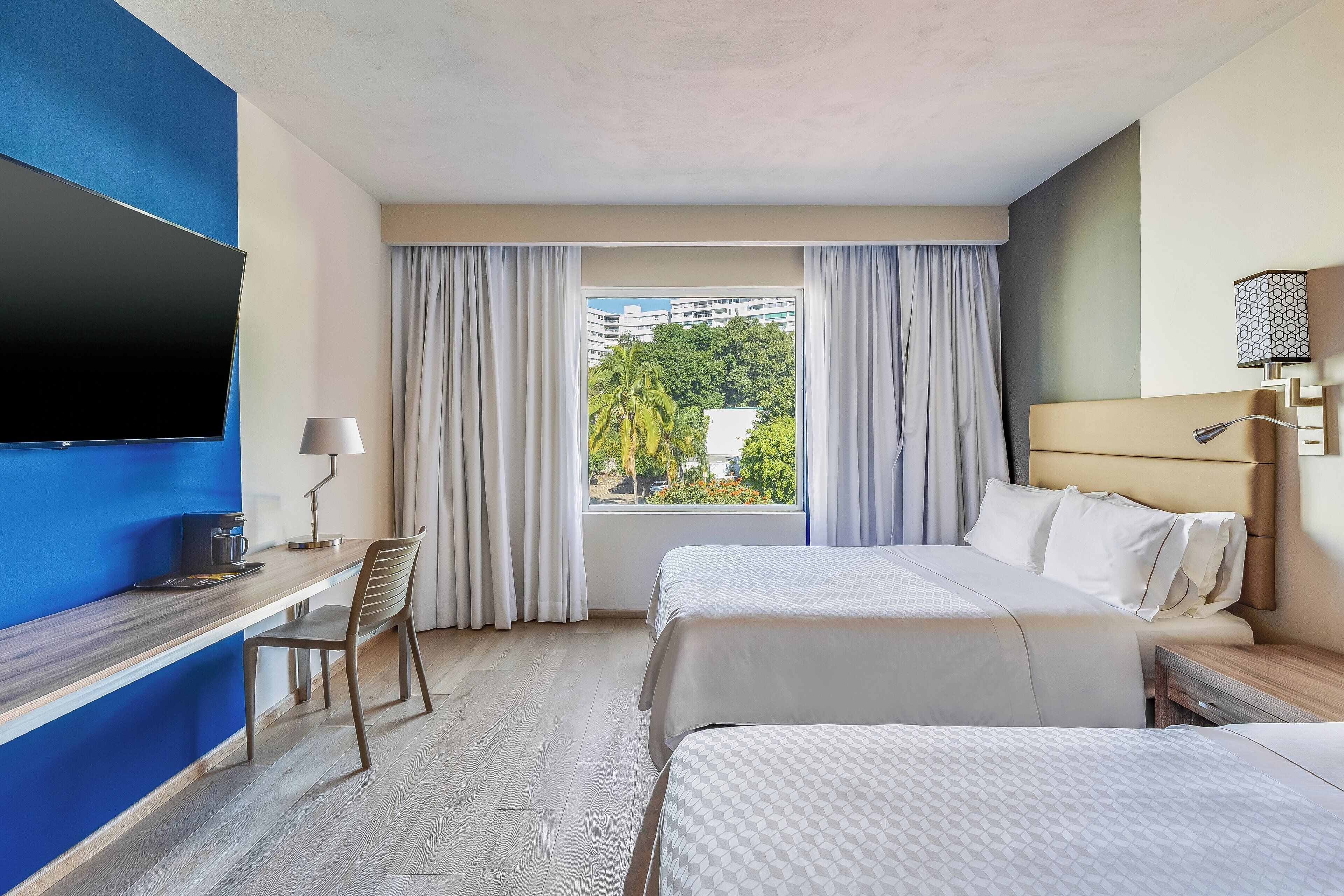 Habitación de hotel con cama, escritorio, silla, televisor y ventana con cortinas.