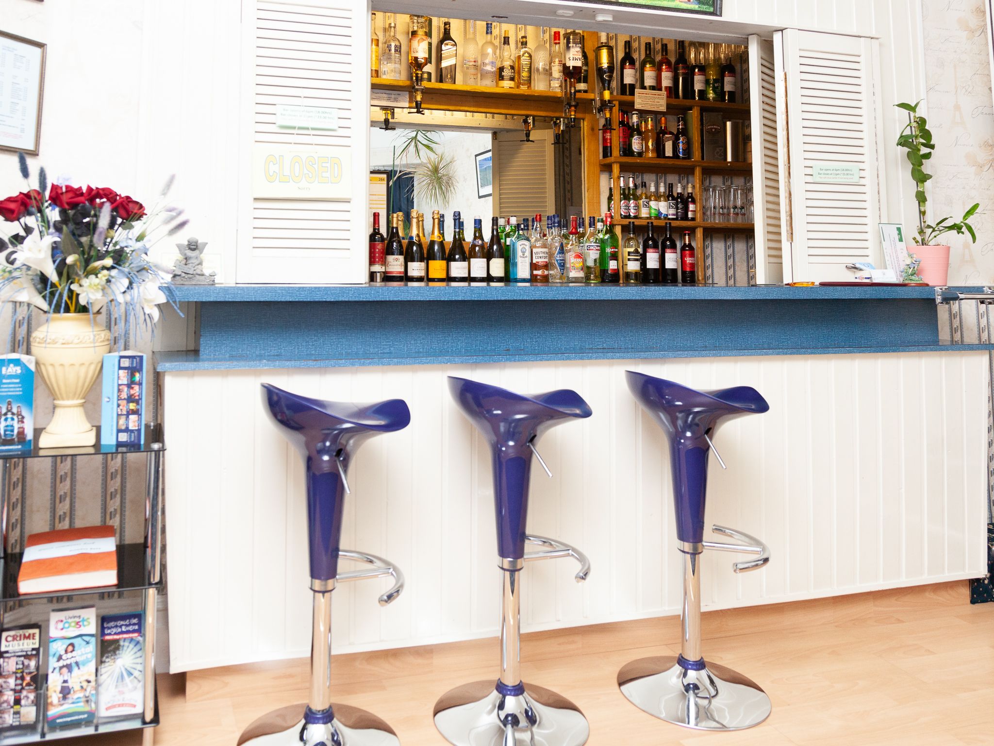A bar area with a blue counter, three blue bar stools, a variety of bottles on shelves, and a 'CLOSED' sign on the wall.