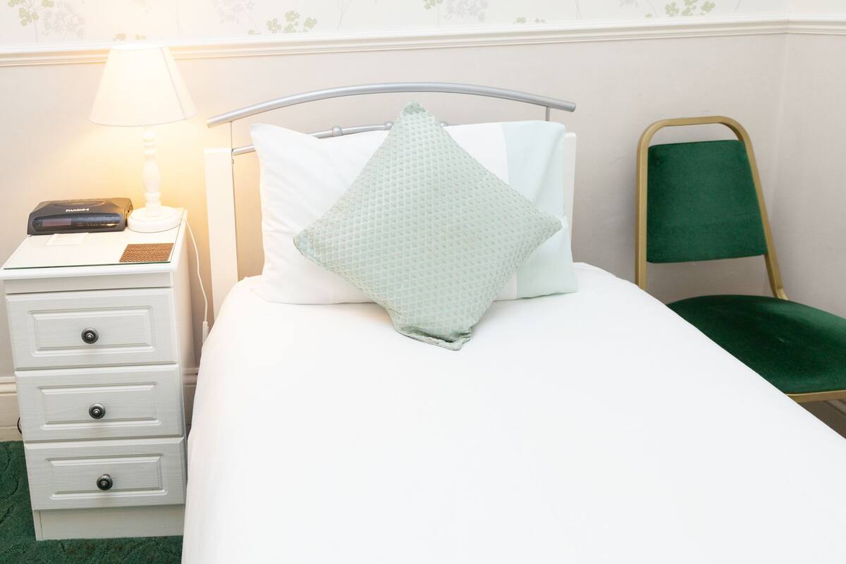 Cama bem arrumada com uma cadeira verde, mesa de cabeceira com abajur e um dispositivo, e parede com papel de parede floral.