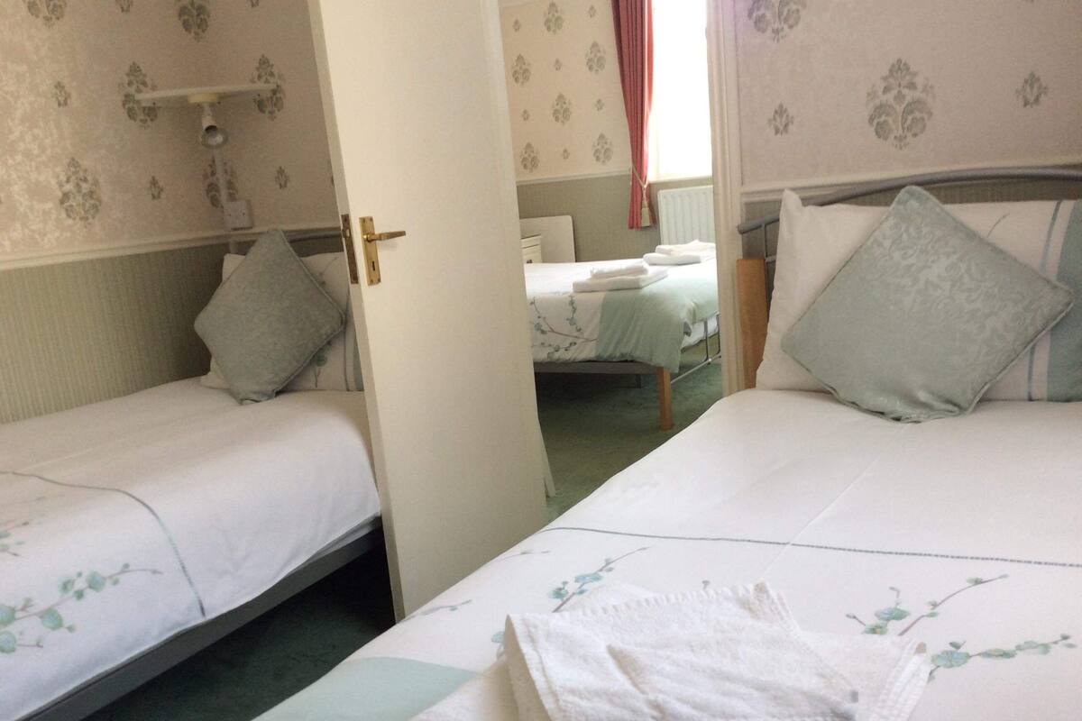 Quarto de hotel com duas camas de solteiro, cada uma com travesseiro e toalha dobrada. O quarto possui papel de parede floral e uma janela com cortinas.