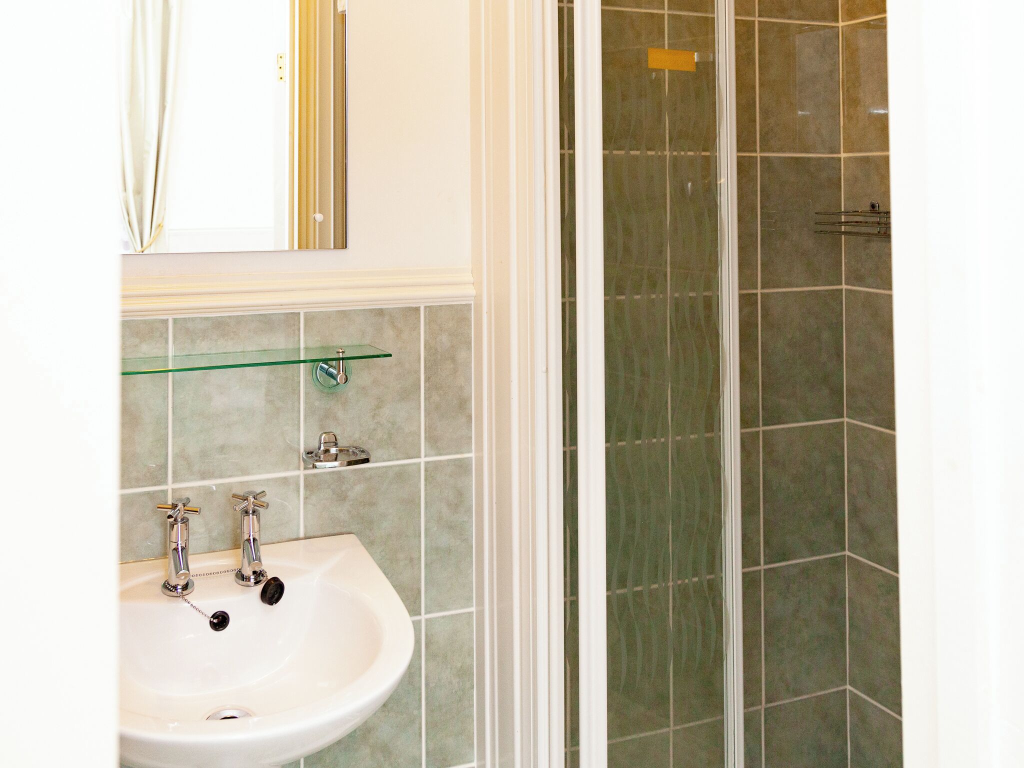 Une salle de bain avec un lavabo blanc, deux robinets, une étagère en verre et un espace douche dont les murs sont recouverts de carreaux.