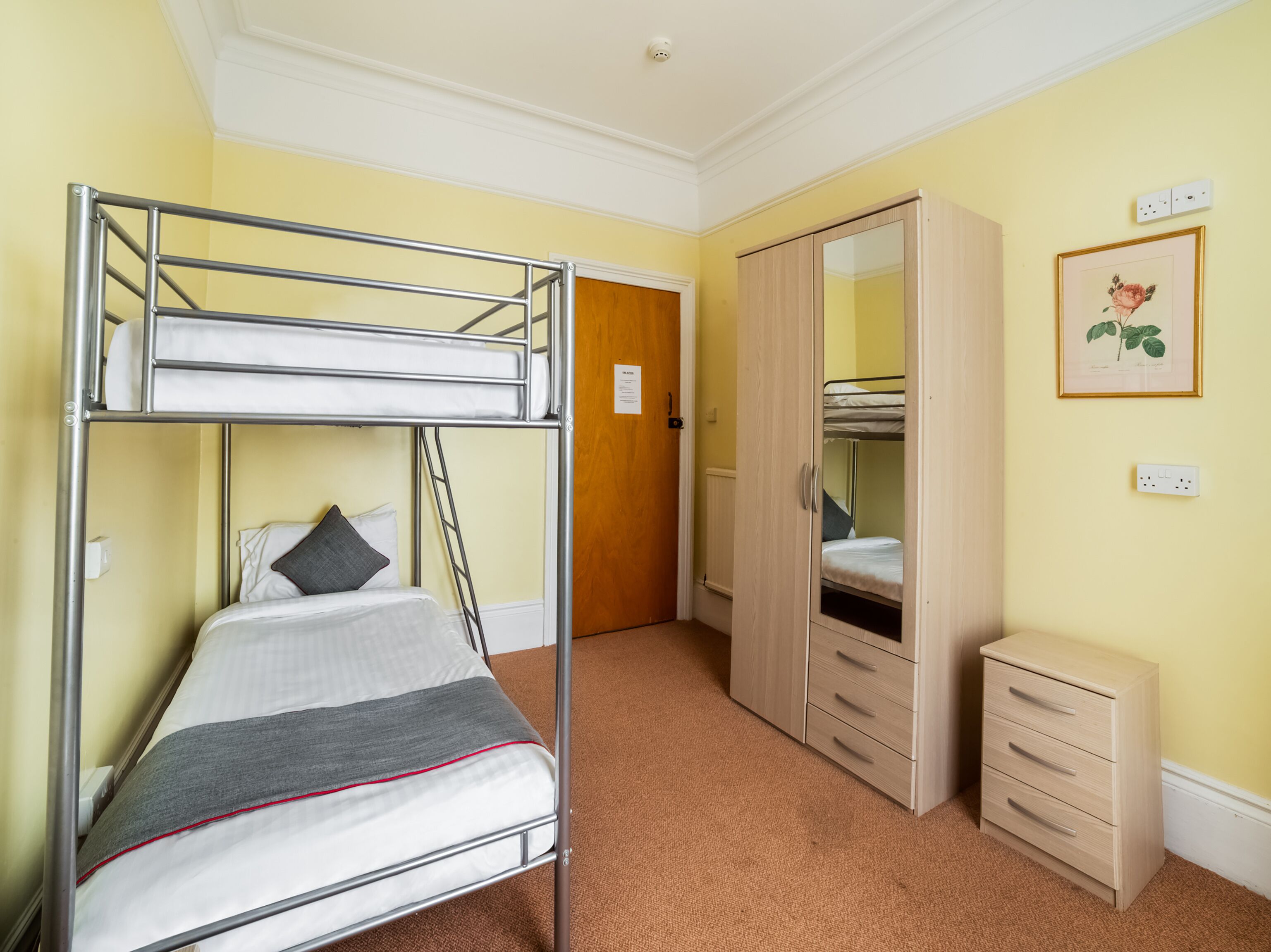 Une chambre avec des lits superposés, une armoire, une table de chevet et une estampe botanique encadrée au mur.