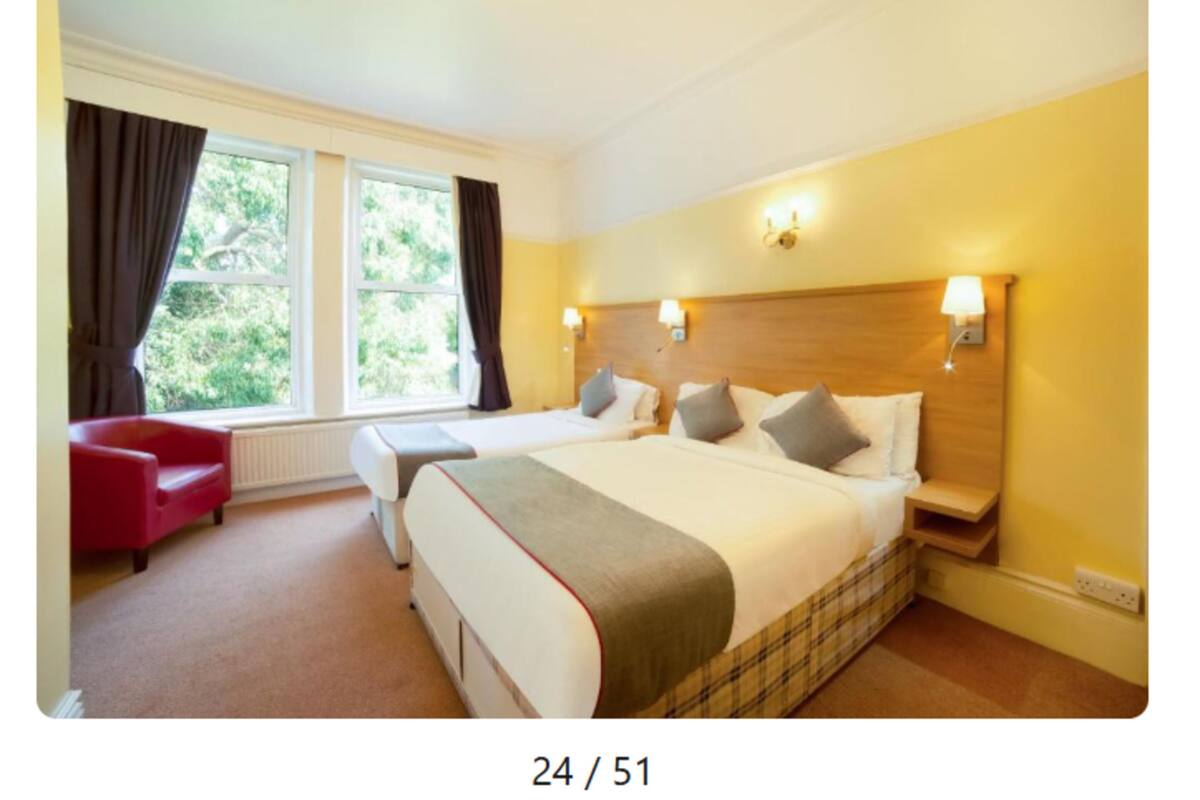 A hotel room with two beds, a red chair, a large window with curtains, and a wooden headboard.