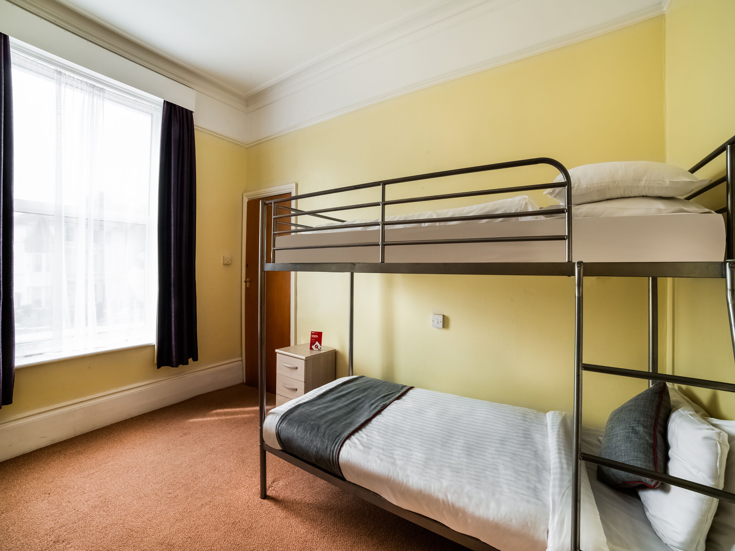 Une chambre avec un lit superposé, une fenêtre, une petite table de chevet et un sol recouvert de moquette.