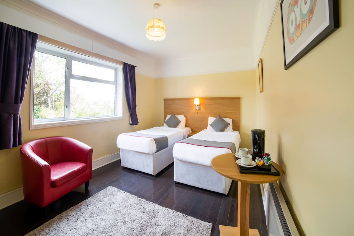 A hotel room with two beds, a red chair, a small table with cups and a tray, and a window with curtains.