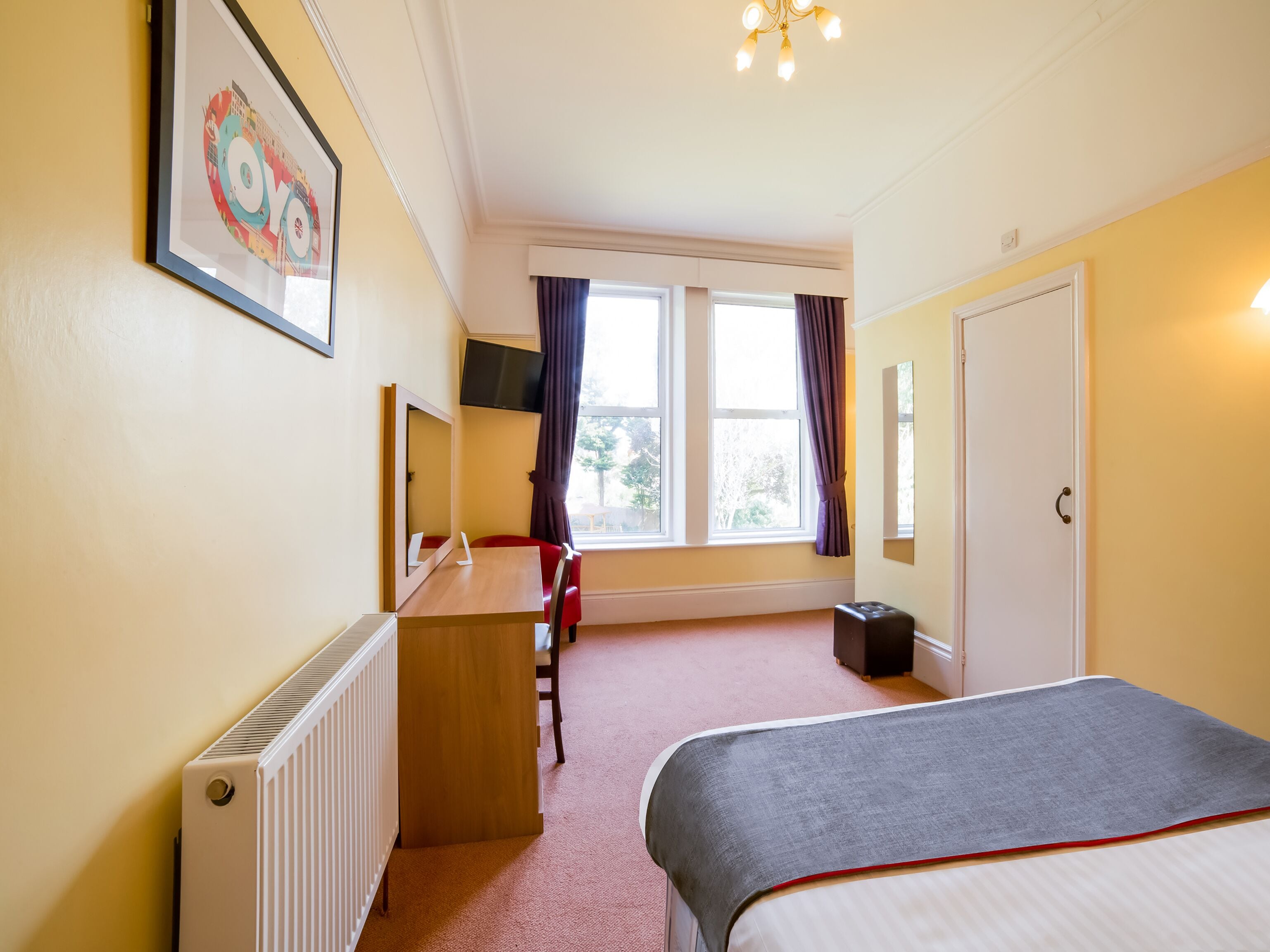 A hotel room with a bed, a desk, a chair, a radiator, a window with curtains, a door, and a ceiling light fixture.