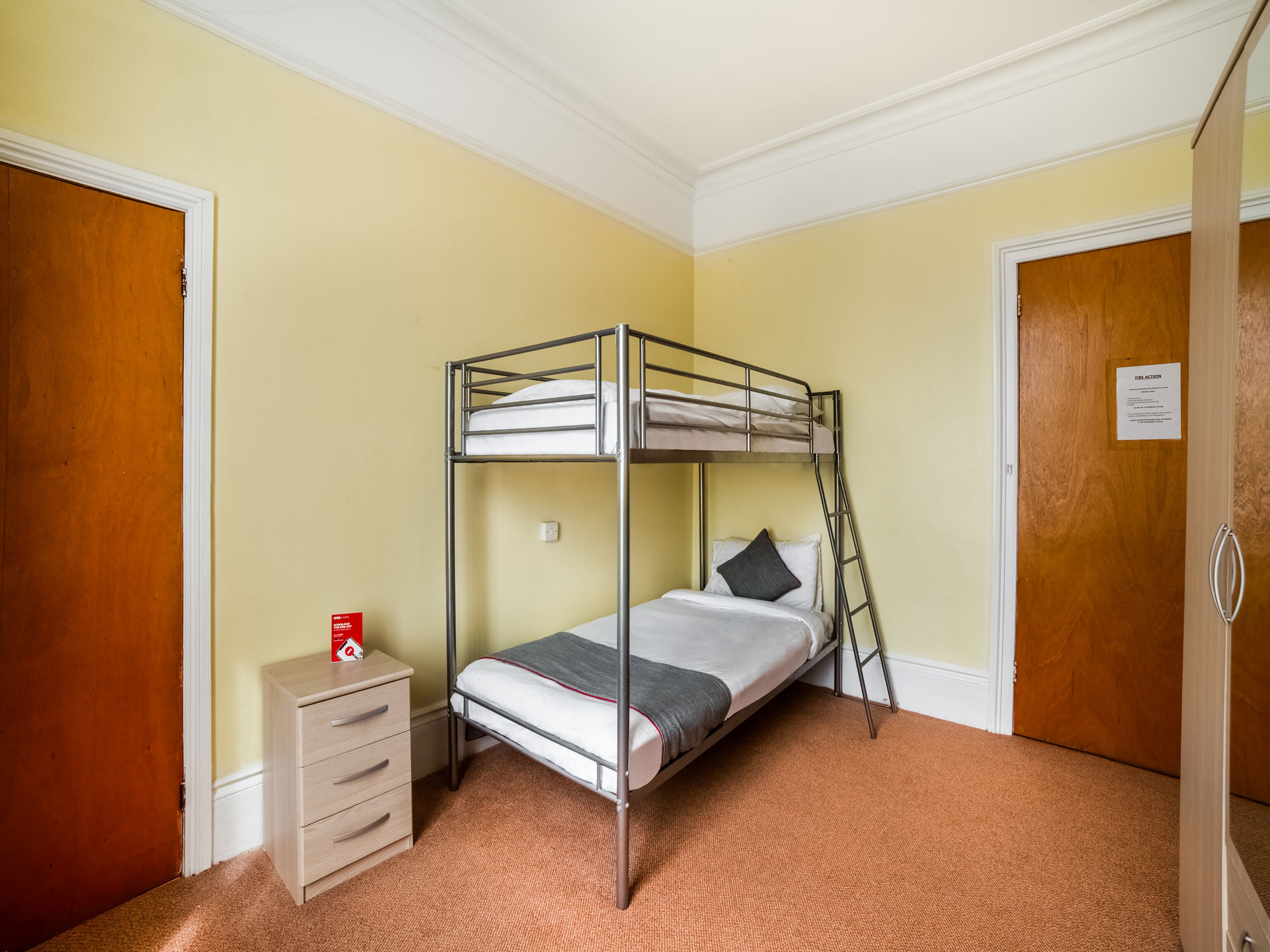 A bunk bed in a room with a wooden door, a small dresser, and a carpeted floor.